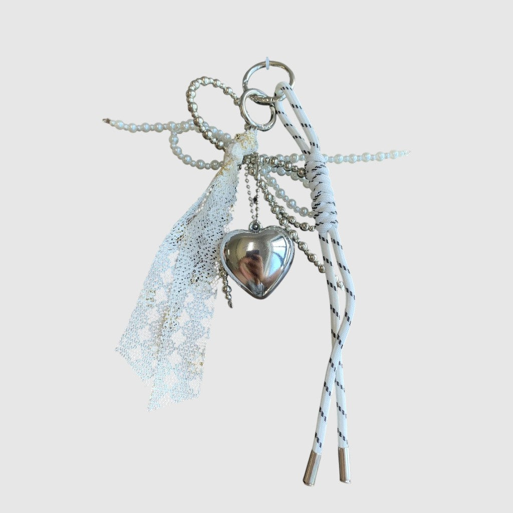 Breloque de sac – Cœur argenté et dentelle. Cœur argenté, dentelle blanche, perles décoratives et cordons blancs à touches noires.