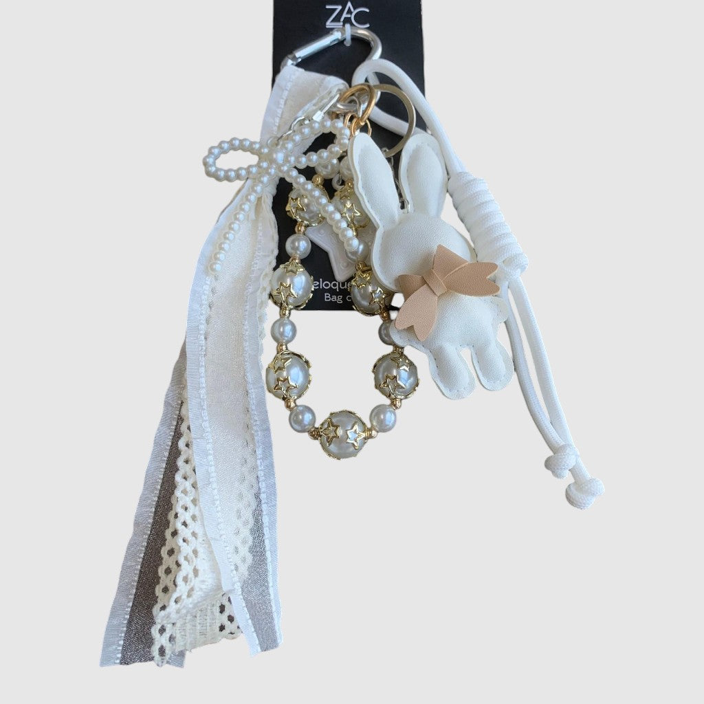 Breloque de sac – Lapin blanc. Lapin blanc à petit nœud beige, perles décoratives ornées d’étoiles dorées, rubans et dentelle.